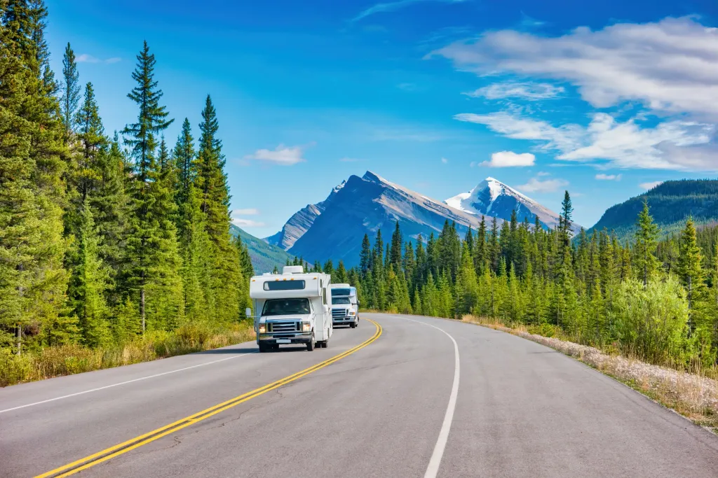 An RV on the road with trees on the side of the road and mountains off in the distance. 